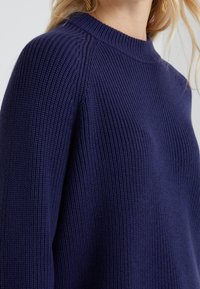 Pull en maille bleu marine avec une texture côtelée, présentant une encolure ras du cou et des coutures d'épaule légèrement abaissées. Design confortable et décontracté.