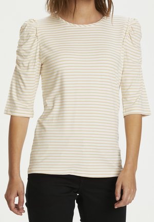 Kobieta w beżowo-białej koszuli w poziome paski z marszczonymi półrękawami i czarnych spodniach, stojąca na białym tle.