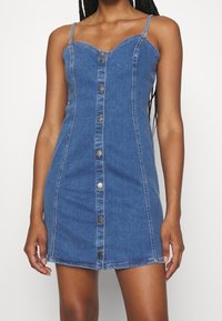 Robe en denim avec une silhouette ajustée, des bretelles spaghetti réglables, des fermetures à boutons à l'avant et un ourlet mi-cuisse. Couleur bleu clair avec des coutures visibles.