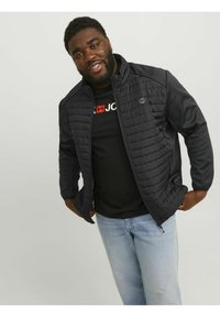 Sort quiltet jakke med et glat stofpanel, fuld lynlås og sidelommer, båret over en sort skjorte og lyseblå jeans.