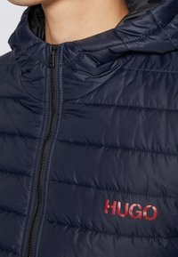 Marineblå dunjakke med hætte, med syninger og en sort lynlås. Logoen "HUGO" i rødt på venstre bryst. Glat tekstur.