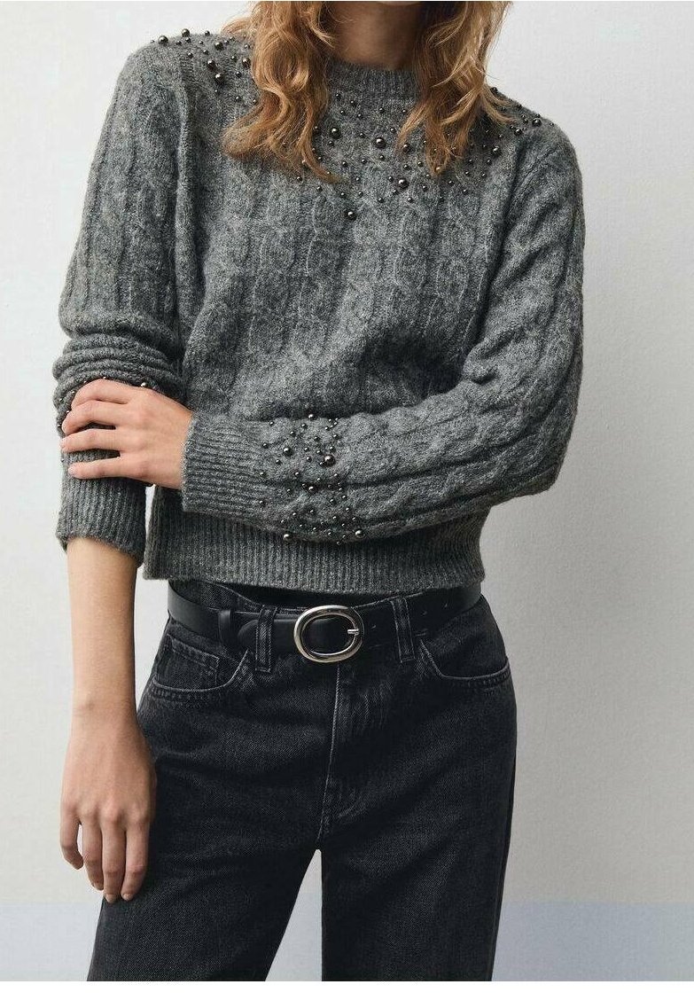 Personne portant un pull en maille gris avec de petites perles sur les épaules et les manches, associé à un jean noir et une ceinture noire avec une boucle argentée.