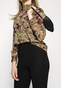 Langärmlige Bluse mit floralem Muster in Grün, Lila und Braun, mit hohem Kragen und gerafften Manschetten, kombiniert mit schwarzen Leggings und einer kleinen Tasche.