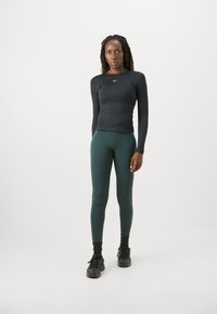 Svart langermet treningsgenser, tettsittende; grønne tights, elastisk stoff; svarte joggesko; minimalistisk design med subtil logo.