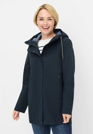 Lächelnde Frau in marineblauer Kapuzenjacke über einem gestreiften Hemd und blauen Jeans, stehend vor einem schlichten weißen Hintergrund.