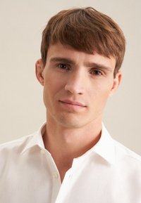 Junger Mann mit kurzen braunen Haaren, der ein weißes Hemd mit Knöpfen trägt und direkt in die Kamera vor einem neutralen Hintergrund blickt.