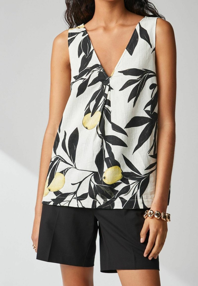 Femme portant un haut blanc sans manches à col en V avec un imprimé feuilles noires et citrons jaunes, associé à un short noir et un bracelet doré.