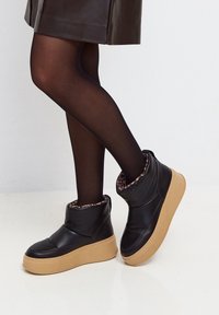 Piernas en medias negras transparentes que llevan botas negras hasta el tobillo con gruesas suelas beiges y forro de estampado animal, combinadas con una falda marrón oscura.