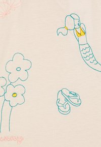 Stof met een roomkleurige achtergrond met kleurrijke borduursels: een zeemeermin in blauw en geel, bloemen in blauw en geel, en slippers in blauw.