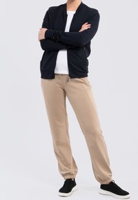 Veste bomber zippée navy, t-shirt blanc en dessous, pantalon jogger beige et baskets noires avec semelles blanches. Tissu doux et coupe décontractée.