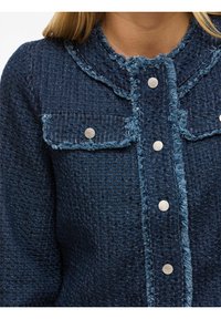 Tamno plavi teksturirani kaput sa pohabanim rubovima, ovratnikom i metalnim dugmadi na prednjem delu, kao i dva džepa na grudima.