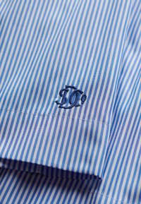 Blåt og hvidt stof med lodrette striber og mørkeblå broderede initialer "DC" nær en søm.