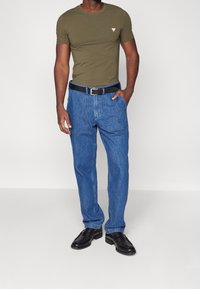 Mand iført olivengrøn T-shirt, blå denimjeans med store forlommer, sort bælte og sorte læder loafers, der står mod en hvid baggrund.