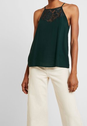 Bezrekawnik ciemnozielony z koronkowym karczkiem, noszony z wysokim stanem i szerokimi białawymi spodniami na stojącej osobie.