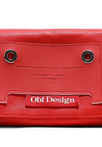 Bolsa de cuero roja con ojales plateados, texto en relieve y una etiqueta de tela con "Obf Design" en blanco. Diseño elegante y minimalista con una textura suave.