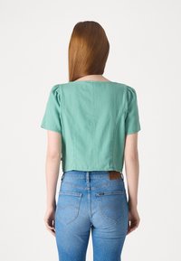 Camicetta corta a maniche corte in tessuto verde menta, con scollatura quadrata e maniche a sbuffo. Abbinata a jeans blu a vita alta.
