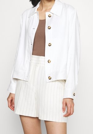 Femme portant une veste blanche boutonnée par-dessus un haut marron sans manches et un short blanc taille haute à fines rayures verticales.