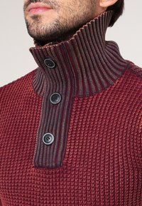 Burgunderfarbener Strickpullover mit hohem Rippkragen, der mit Knopfdetails versehen ist, und einem strukturierten Muster auf dem Körper für visuelles Interesse.