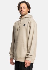 Beige hoodie gemaakt van zachte stof, met een frontzak, geribde manchetten en een klein zwart logo-opdruk op de borst.