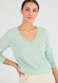 Pull en maille vert menthe clair, design en V, manches longues, texture douce, poignets côtelés, porté avec un pantalon de couleur claire.