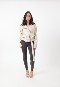 Beige motorjack van kunstleer met een ceintuur in de taille, ritssluiting en brede kraag, gecombineerd met zwarte slim-fit broek en hoekige hakken.