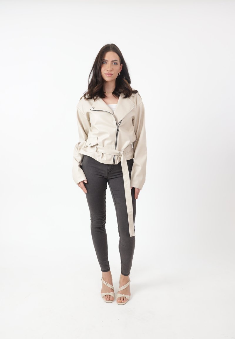Giacca in faux leather beige chiaro con vita cinturata, zip asimmetrica e colletto ampio, abbinata a jeans skinny neri e tacchi nudi.