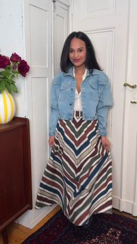 Mujer con cabello largo y oscuro que lleva una chaqueta vaquera azul claro y una falda larga multicolor con rayas en forma de chevron, de pie en el interior cerca de un jarrón con flores.