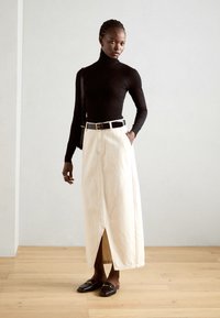 Zwarte coltrui, crèmekleurige maxi-jurk met een voorste splijt, gecombineerd met een zwarte riem en bruine loafers met een gouden accent. Soepele texturen.