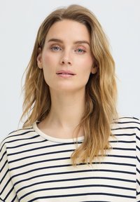 Jeune femme aux longs cheveux blonds portant un t-shirt rayé blanc et marine, regardant calmement l'appareil photo sur un fond clair uni.