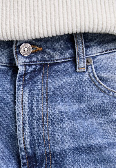 Világoskék farmernadrág gombos záródással és kontrasztos narancssárga varrással, látható textúrával és közeli felvételen a derékrész részleteivel.