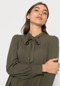 Blusa verde-azeitona com colarinho alto, detalhe de laço e mangas longas. Apresenta ombros franzidos e acentos pregueados ao longo da yoke.