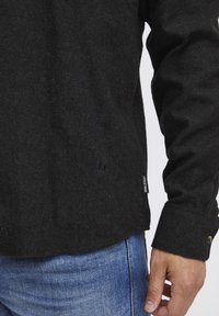 Camisa negra de manga larga en tejido texturizado con un sutil detalle de logo en el dobladillo. Cuenta con un puño abotonado y un corte relajado.