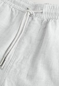 Pantalons gris clair avec une taille élastique et un cordon de serrage avec des embouts en métal, fabriqués à partir d'un tissu texturé. Vue rapprochée des détails.