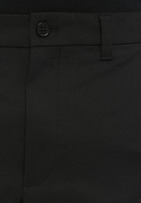 Gros plan de la taille d’un pantalon noir montrant un bouton, une passants de ceinture et une partie de la couture de la poche avant.