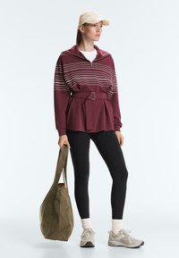 Femeie purtând șapcă bej, pulover cu fermoar bordo cu centură și dungi albe, colanți negri, șosete albe, adidași gri, ținând o geantă tote verde măsliniu.