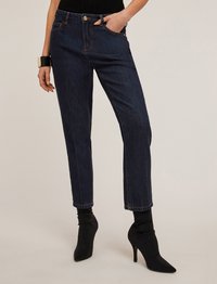 Pantalones de mezclilla azul oscuro con corte slim, que cuentan con un cierre de botón, bolsillos inclinados y puños enrollados. Combinados con botas de tacón puntiagudas negras.