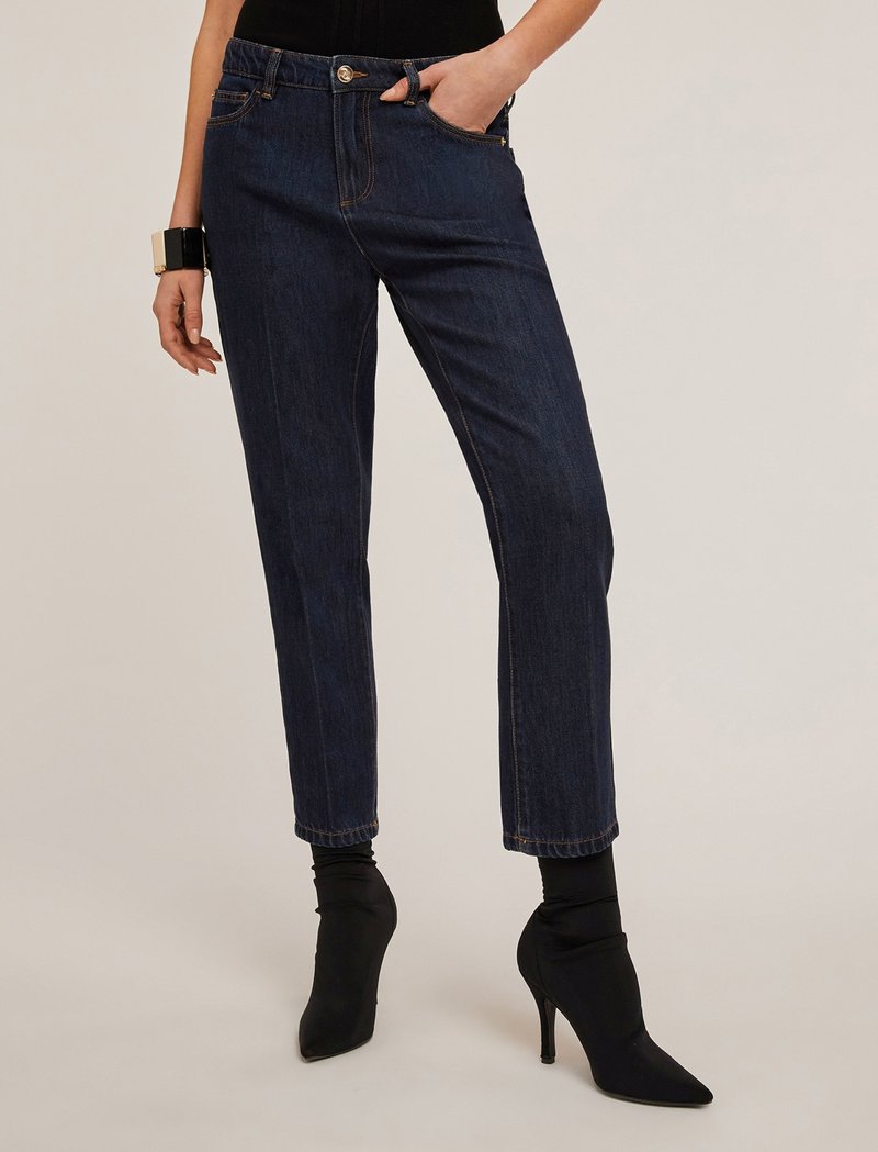 Pantalones de mezclilla azul oscuro con corte slim, que cuentan con un cierre de botón, bolsillos inclinados y puños enrollados. Combinados con botas de tacón puntiagudas negras.