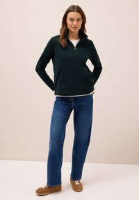 Fleece zippé vert foncé avec un col haut, doté d'une poche frontale, associé à un jean bleu et des baskets marron. Texture lisse, coupe décontractée.