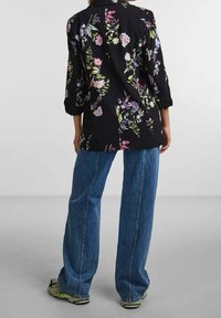 Personne portant un jean bleu en denim et un blazer noir à fleurs, debout sur un sol gris devant un mur blanc uni.