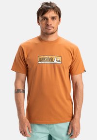 Camiseta de manga corta naranja de algodón, con un logotipo de Quiksilver y un gráfico de montaña y ola en blanco y negro. Textura suave, cuello redondo.