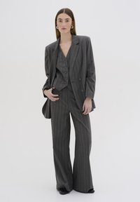 Abito grigio a righe sottili con blazer doppiopetto, gilet su misura con bottoni e pantaloni a gamba larga. Texture liscia e design strutturato.
