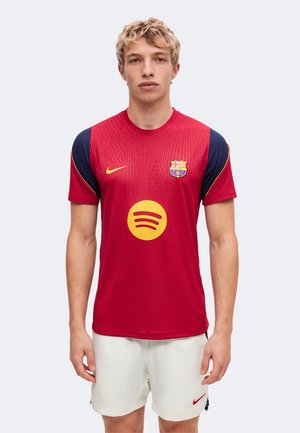 Jeune homme portant un maillot de football rouge et bleu marine du FC Barcelone avec des logos jaunes Nike et Spotify et un short blanc Nike, debout devant un fond uni.