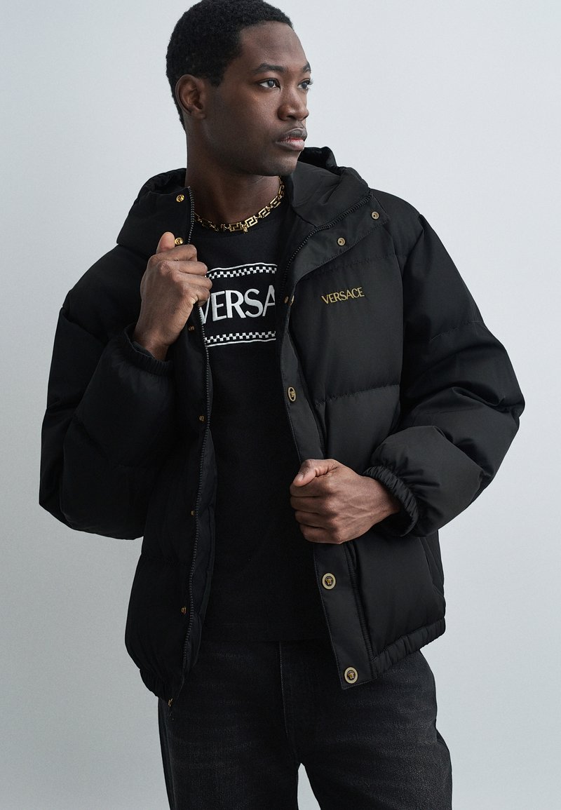 Melna pūkainā jaka ar kapuci, ar zelta aksesuāriem un logo. Mīksta tekstūra, elastīgas aproces un brīva piegriezuma. Uzvilkta virs melna T-krekla.