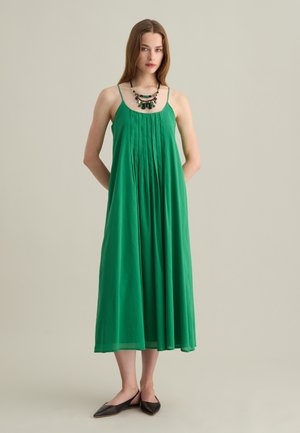 Abito maxi verde con spalline sottili, design plissettato, tessuto morbido e silhouette fluida. Abbinato a una collana multicolore e scarpe basse.