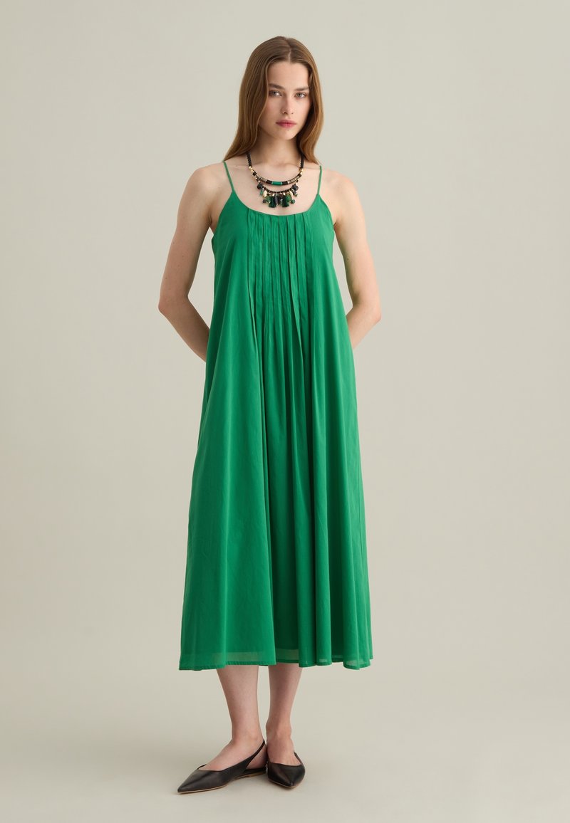 Abito maxi verde con spalline sottili, design plissettato, tessuto morbido e silhouette fluida. Abbinato a una collana multicolore e scarpe basse.