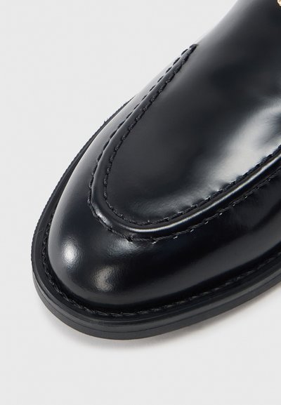 Mocasín de cuero negro pulido con costuras detalladas en la parte superior y puntera redondeada sobre un fondo claro y simple.