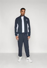 Σκούρο μπλε φόρμα με ζακέτα με φερμουάρ και λευκές πλαϊνές λωρίδες, καθώς και παντελόνια που ταιριάζουν. Το ύφασμα είναι λείο· λεπτομέρειες περιλαμβάνουν μικρό λογότυπο στη ζακέτα.