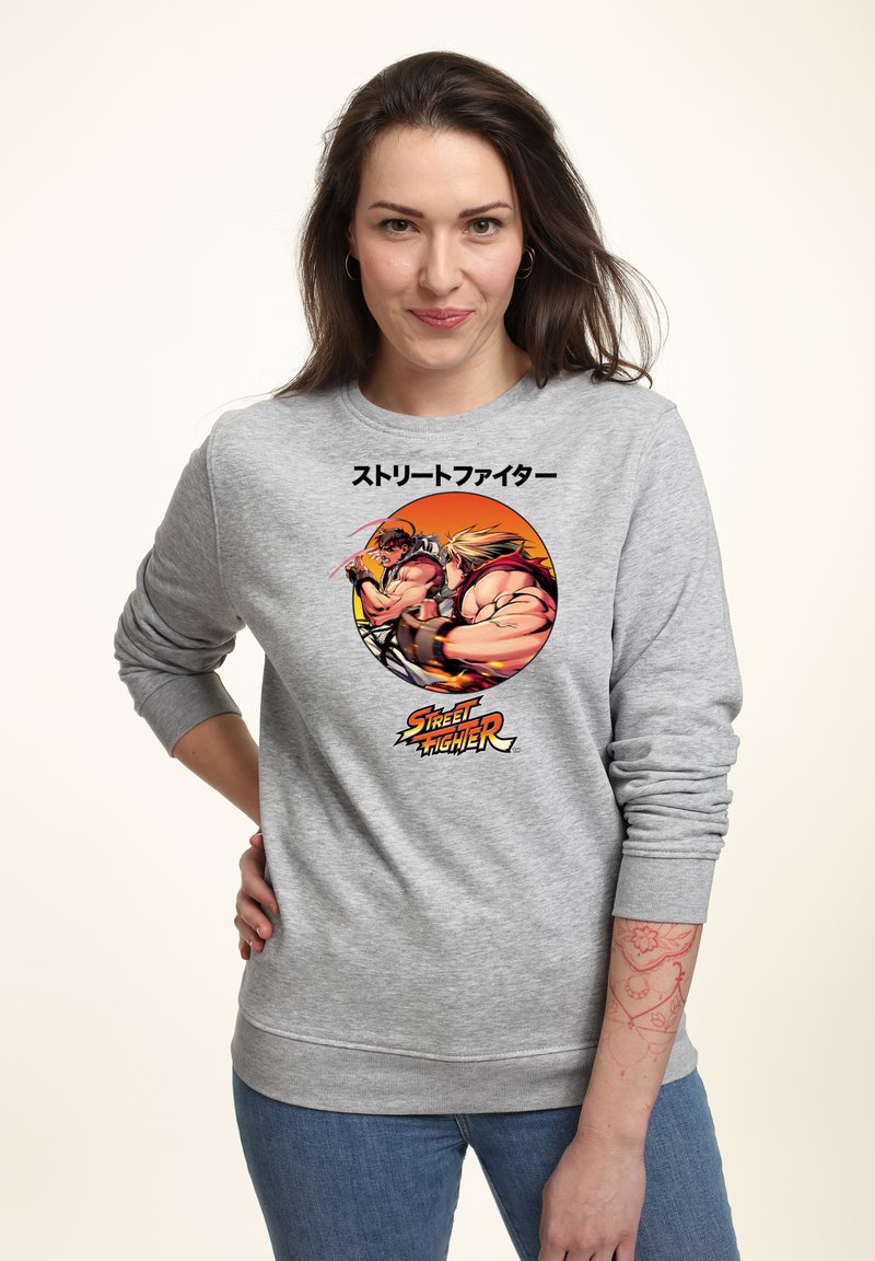 Vrouw draagt een grijze sweatshirt met kleurrijke Street Fighter-personages en Japanse tekst, staand met een hand op haar heup tegen een lichte achtergrond.