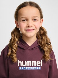 Smilende ung pige med lysbrunt krøllet hår sat op i hestehaler iført en vinrød "hummel SPORTSWEAR" hættetrøje mod en ensfarvet lys baggrund.