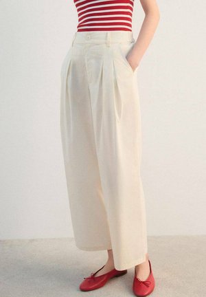 Pantalones anchos de pierna ancha color crema con pliegues delanteros y bolsillos laterales, confeccionados en tela ligera, combinados con zapatos planos rojos. Se ve una blusa de rayas.